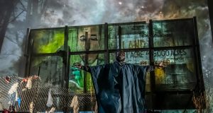 un comédien bras écartés, devant un panneau publicitaire délabré, et en arrière plan, une forêt mystérieuse et un ciel étoilé