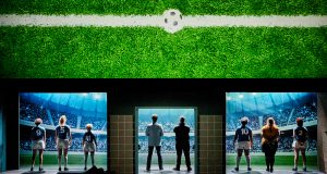 équipe féminine de football, de dos, face au stade. Au dessus d'elles, un grand écran projetant un ballon de football sur du gazon.