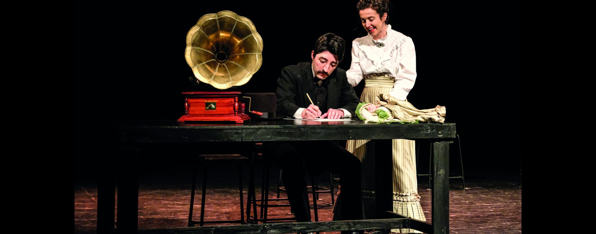 deux personnages en costumes d'époque écrivent à une table sur laquelle repose un grammaphone